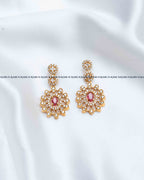 A pair of gold-toned Indian-style dangle earrings with a central pink stone and clear rhinestone floral detailing, displayed on a white satin fabric background.