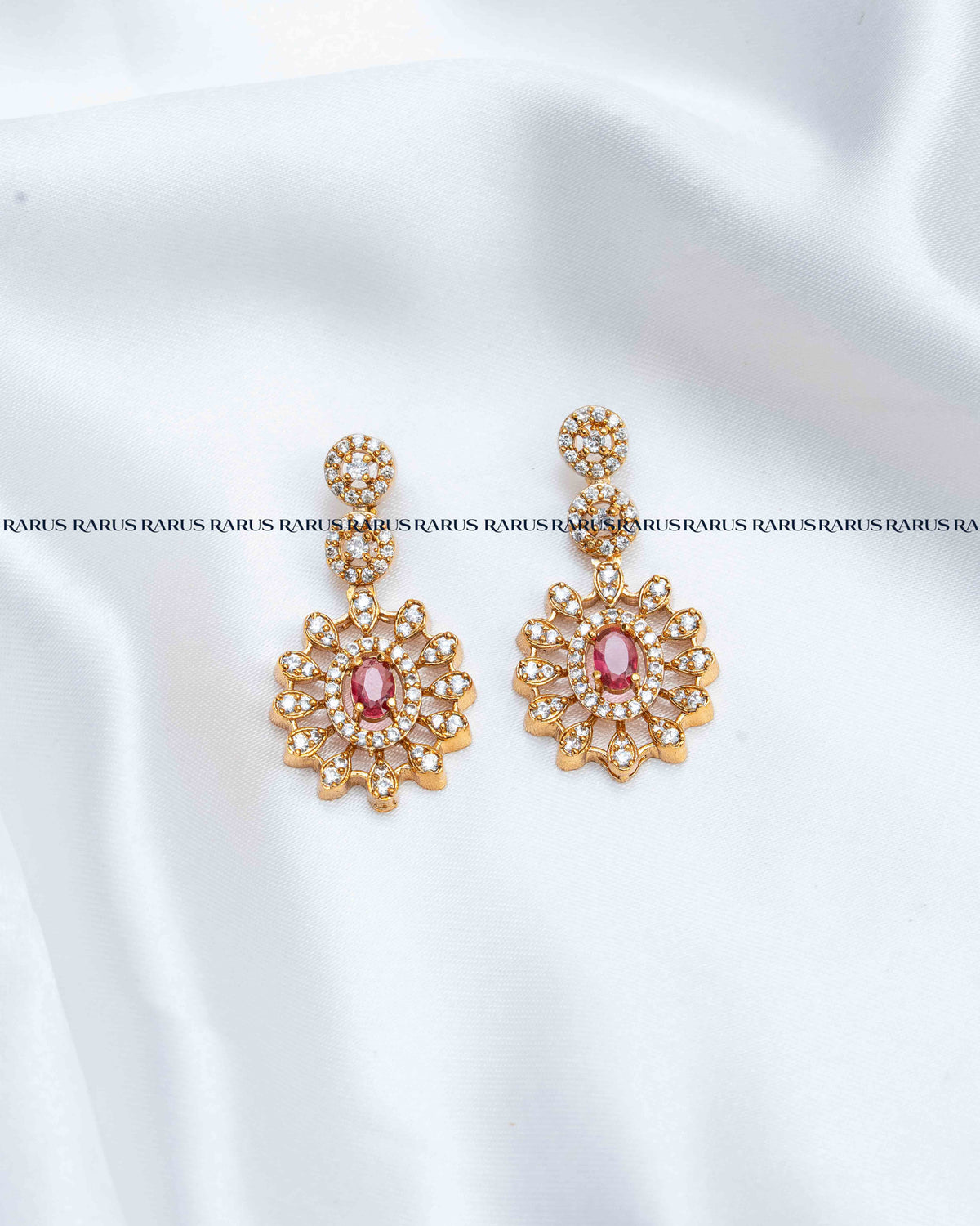 A pair of gold-toned Indian-style dangle earrings with a central pink stone and clear rhinestone floral detailing, displayed on a white satin fabric background.