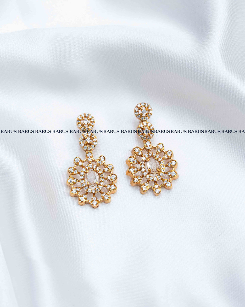 A pair of gold-toned statement earrings with intricate floral crystal designs, displayed on a white satin fabric background.