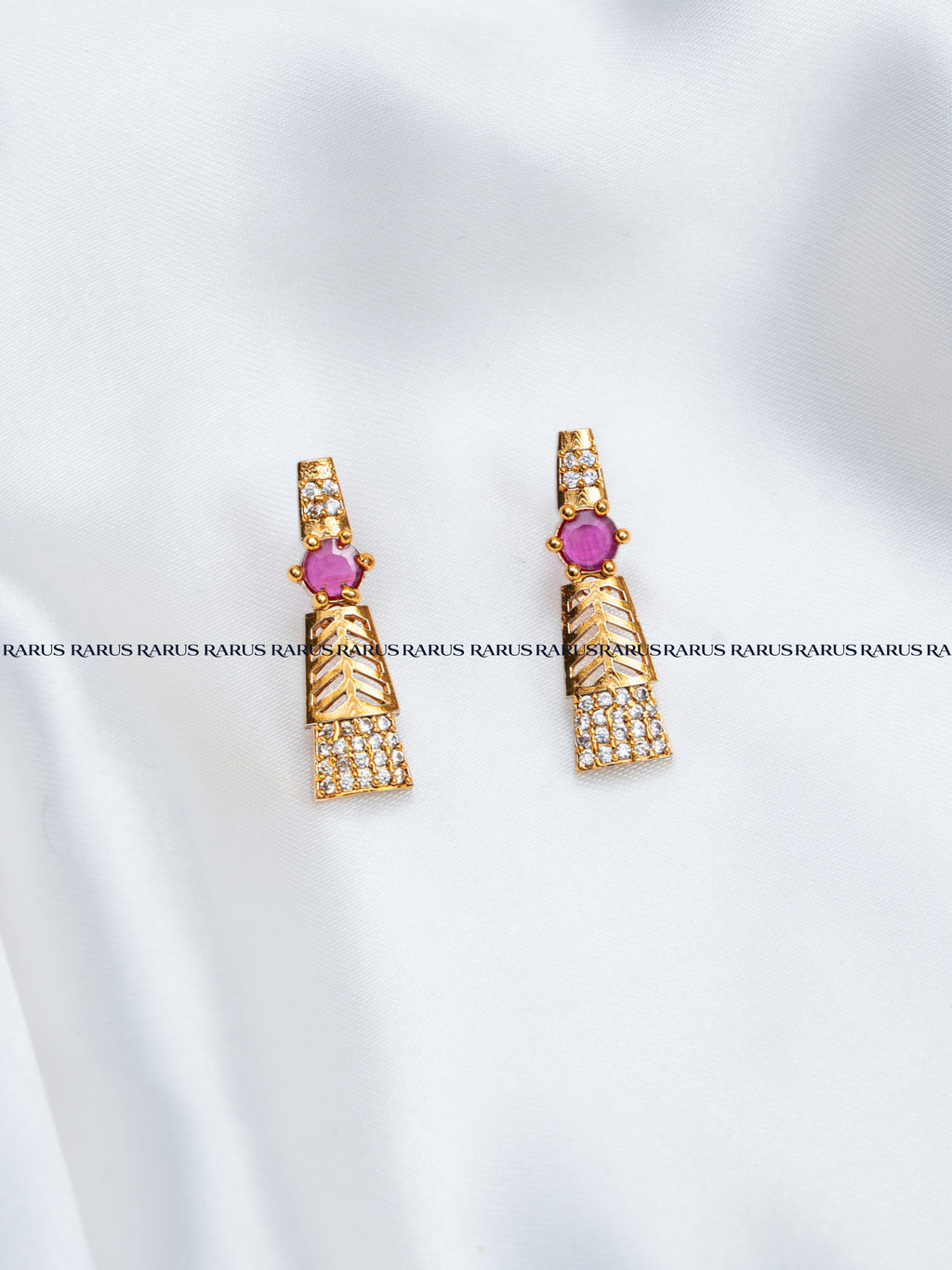 A pair of gold-toned geometric drop earrings with a central round pink gemstone and rows of small clear stones, displayed on a white fabric background with a repeating watermark.