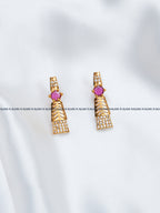 A pair of gold-toned geometric drop earrings with a central round pink gemstone and rows of small clear stones, displayed on a white fabric background with a repeating watermark.