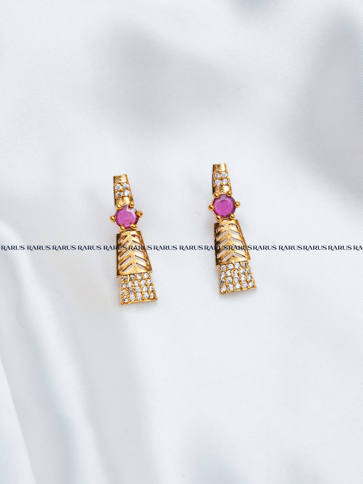 A pair of gold-toned geometric drop earrings with a central round pink gemstone and rows of small clear stones, displayed on a white fabric background with a repeating watermark.