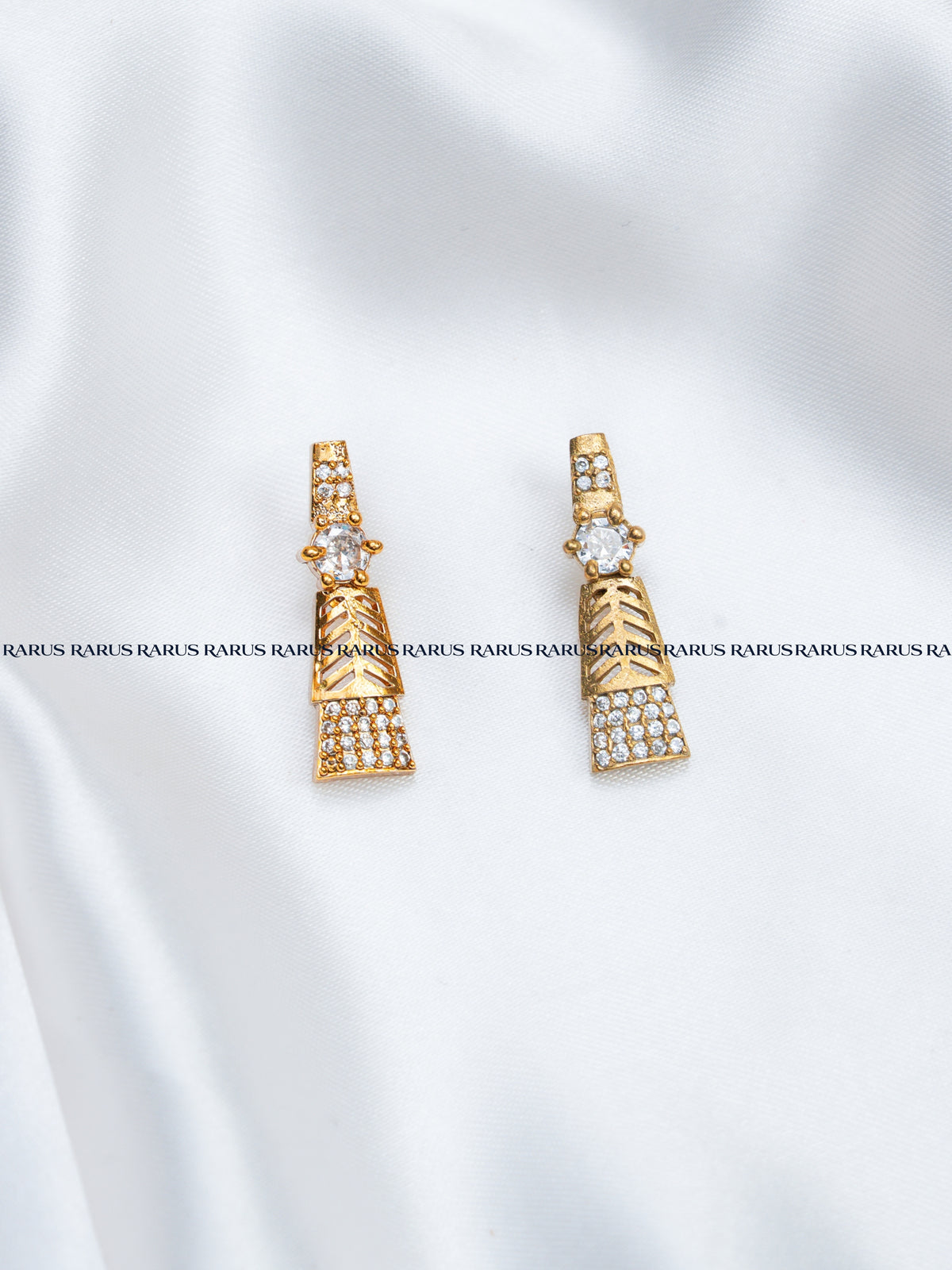A pair of gold-toned statement earrings with clear rhinestone detailing, displayed on a white satin fabric background with a repeating watermark.