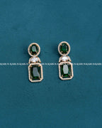 A pair of gold drop earrings with green gemstones, displayed on a textured teal background. The earrings feature an oval top with a green stone surrounded by small clear stones, and a larger rectangular green stone below, separated by a small clear stone.