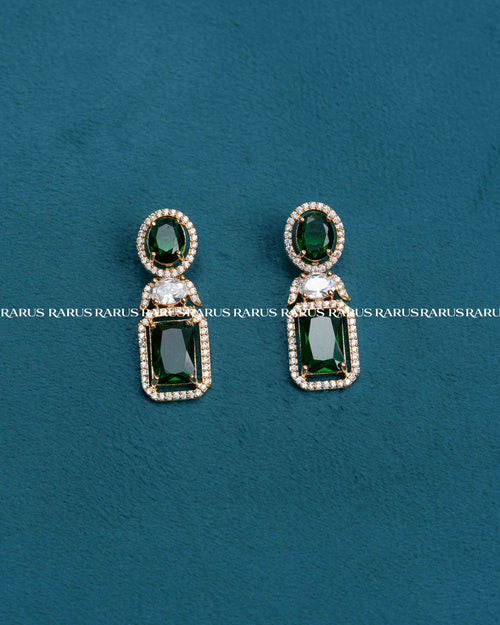 A pair of gold drop earrings with green gemstones, displayed on a textured teal background. The earrings feature an oval top with a green stone surrounded by small clear stones, and a larger rectangular green stone below, separated by a small clear stone.