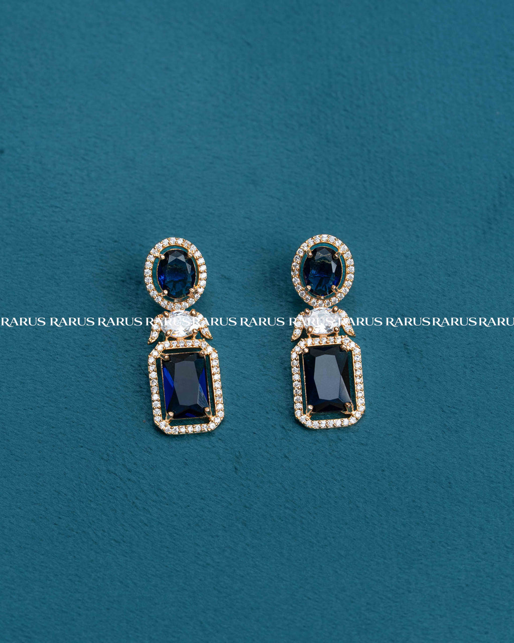 A pair of gold drop earrings with dark blue rectangular and oval stones, accented by small clear crystals, displayed on a textured teal background.