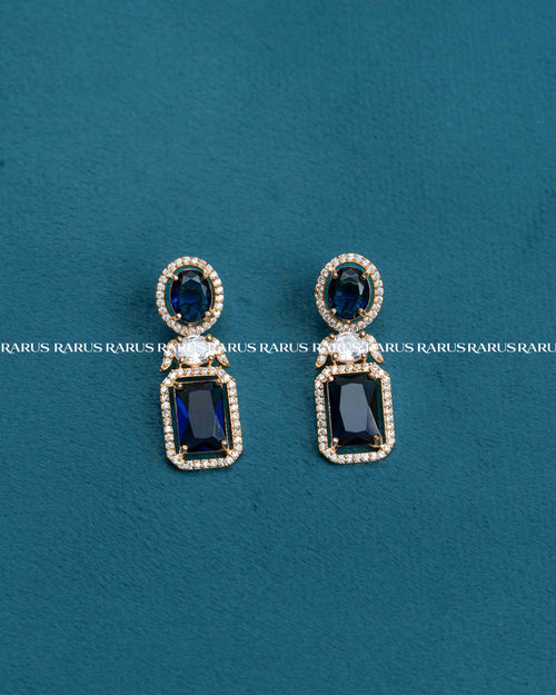 A pair of gold drop earrings with dark blue rectangular and oval stones, accented by small clear crystals, displayed on a textured teal background.