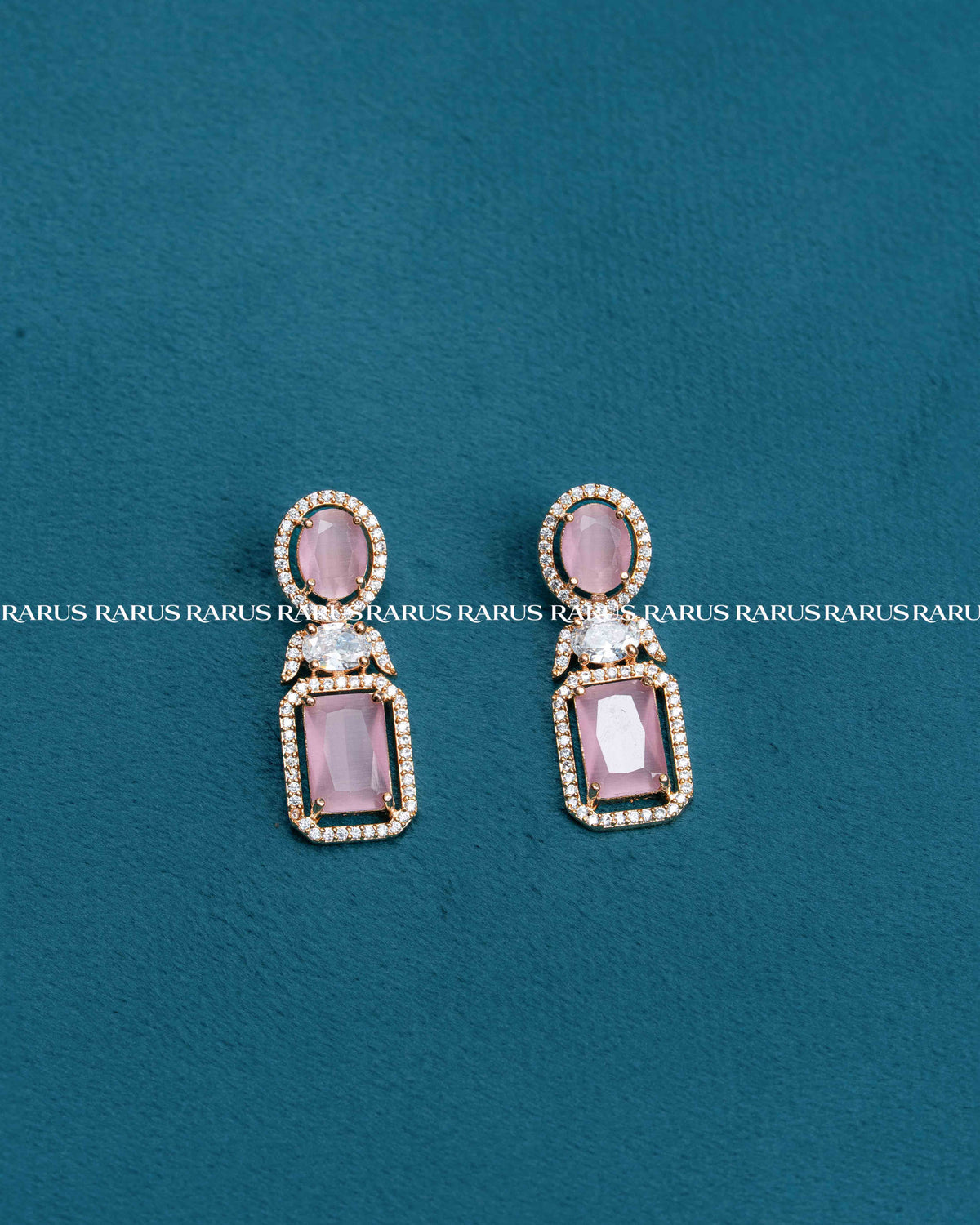 A pair of gold drop earrings with oval pink gemstones and rectangular pink gemstones, accented with small clear stones, displayed on a textured teal background.