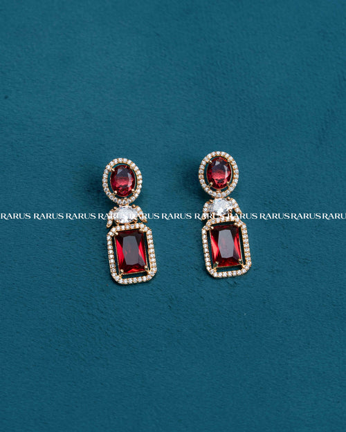 A pair of gold-plated drop earrings featuring oval and rectangular deep red gemstones surrounded by small clear crystals, displayed on a textured dark teal background.
