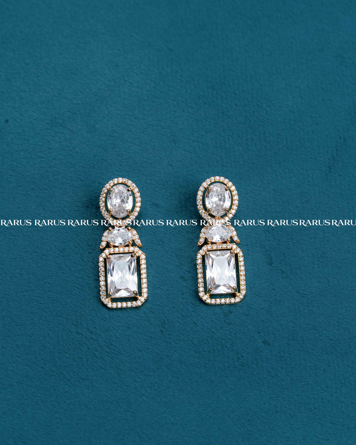 A pair of gold drop earrings with clear rectangular and oval gemstones, set against a textured teal background with a repeating watermark.
