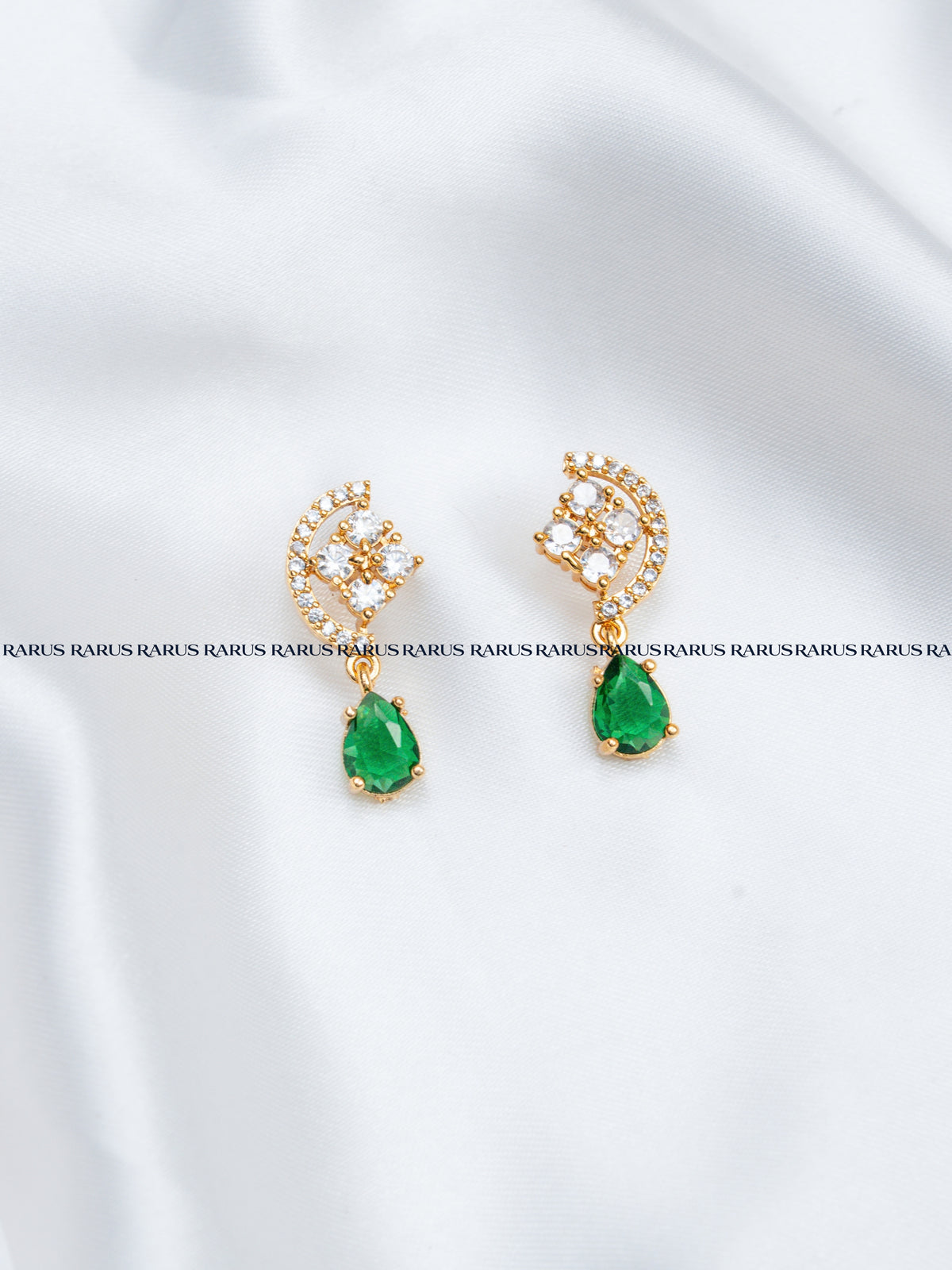 A pair of gold crescent-shaped drop earrings with clear rhinestones and green teardrop gemstones, displayed on a white satin fabric background.