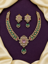 An ornate gold jewelry set with a necklace and matching earrings, featuring green stones, pink center stones, and white stones, arranged on a dark purple background with cream satin draping.